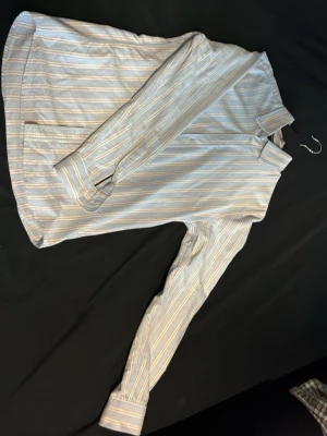 Randig skjorta från Bondelid, S - Snygg skjorta från Bondelid i ljusblått med tunna vita och beige ränder. Klassisk krage, långa ärmar och knäppning framtill. Skjortan har en skräddad passform och är tillverkad i mjuk bomull, perfekt för en clean och stilren look. Säljs då den sällan kommer till användning.