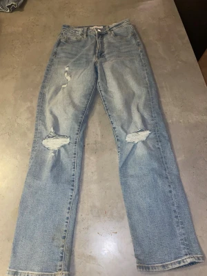 Ljusblå ripped jeans med straight fit - Säljer ett par ljusblå jeans med snygga slitningar på knäna och låret. Modellen är högmidjad med rak passform och klassiska fem fickor. Jeansen har en cool vintagekänsla och är perfekta för dig som gillar en avslappnad streetstil.