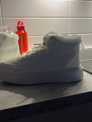 Vita sneakers med fuskpäls och plattform - Säljer ett par vita sneakers med högt skaft och fluffig fuskpäls runt kanten. Skorna har snörning, platt sula med plattform och är tillverkade i syntetmaterial med skinnliknande yta. Perfekta för dig som vill ha en clean look med extra cozy vibe.