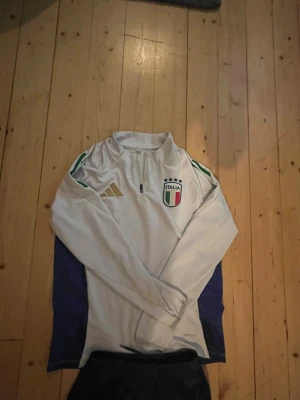 Vit Adidas Italia half zip träningströja - Säljer en snygg vit långärmad träningströja.Med Italien och adidas. OBS ena dragkedjan är borta på vänstra ben.