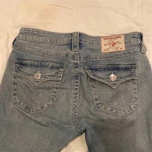 True Religion Becca Bootcut Jeans - Snygga ljusblå bootcut jeans från True Religion, modell Becca Mid Rise Bootcut. Jeansen har klassiska fem fickor, coola detaljerade bakfickor med lock och silverknappar samt tydliga sömmar. Tillverkade i mjukt denim med lätt slitning framtill.