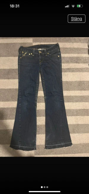 True Religion bootcut jeans med guld - Säljer ett par mörkblå bootcut jeans från True Religion med ikoniska guldiga detaljer och broderier på fickorna. Jeansen har låg midja, slitna benslut och snygga kontrastsömmar. 