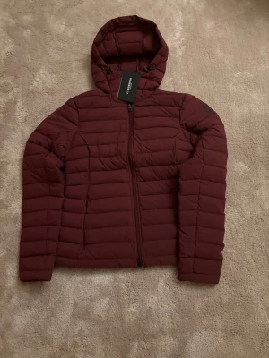 Vinröd pufferjacka med huva - Säljer en vinröd pufferjacka från Lindbergh med huva och dragkedja framtill. Jackan har quiltad design och två sidofickor med dragkedja. Perfekt för kyliga dagar och har en snygg, modern look.