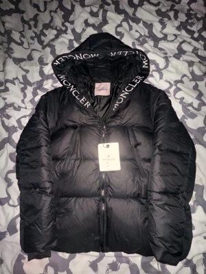 Svart Moncler pufferjacka med huva - Svart pufferjacka från Moncler i storlek M. Jackan har en stor huva med Moncler-logga, två dragkedjeförsedda fickor och en snygg Moncler-patch på ärmen. Materialet är glansigt och jackan har en kraftig dragkedja framtill. Perfekt för kalla dagar.