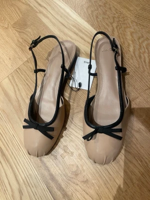 Beige ballerinaskor med svarta detaljer - Snygga ballerinaskor från Mango i beige med svarta remmar och rosett framtill. Skorna har rund tå, platt sula och är tillverkade i syntetmaterial (polyuretan). Perfekt för dig som gillar stilrena och trendiga detaljer.