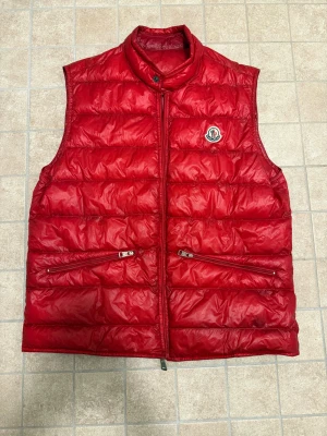 Röd dunväst från Moncler - Säljer en snygg röd dunväst från Moncler med klassisk logga på bröstet. Västen har ståkrage, två dragkedjefickor och quiltad design. Perfekt för lager-på-lager och har en modern, sportig look. Dragkedja framtill och detaljer i mörkröd ton.