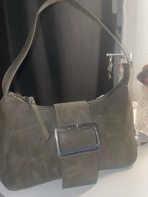 Grå axelväska med stor spänne - Snygg grå grön axelväska i skinnimitation med bred axelrem och ett stort metallspänne framtill. Väskan har dragkedja upptill och en extra ficka med dragkedja på baksidan. 