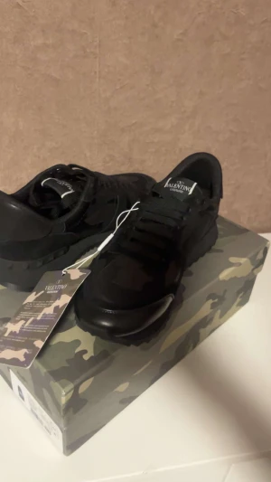 Valentino Garavani Rockrunner sneakers svart - Svarta Valentino Garavani Rockrunner sneakers med coolt kamouflagemönster. Snygga detaljer i mocka och skinn, grov gummisula med pyramidmönster och rund tå. Klassisk snörning och logga på plösen. Perfekt för dig som vill ha en exklusiv streetstil.