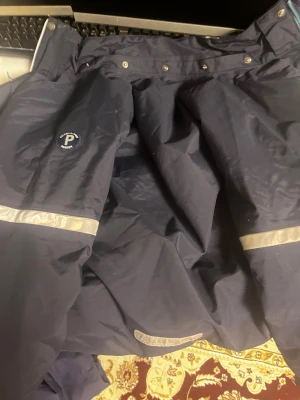 Mörkblå täckbyxor Polarn O. Pyret 146 - Täckbyxor från Polarn O. Pyret i mörkblått med ljusblå fleecefoder. Vattentäta, vindtäta och med reflexdetaljer runt benen. Dragkedja framtill och elastisk midja. Perfekta för vintern och utomhusaktiviteter.