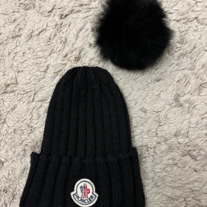 Svart stickad mössa med pälsboll - Mössa med pälsboll, den kan knytas till mössan med ett snöre. Den är använd så den är lite luddig, tvättad är den också 