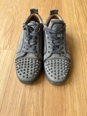 Grå Christian Louboutin sneakers med nitar - Säljer ett par grå sneakers från Christian Louboutin med coola nitar på tån och klassisk röd sula. Skorna är gjorda i mocka med skinnfoder och har snörning framtill. Perfekt för dig som vill sticka ut med en edgy och lyxig stil.