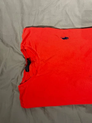 Orange t-shirt från Polo Ralph Lauren - Snygg orange t-shirt från Polo Ralph Lauren med klassisk blå logga broderad på bröstet. Modellen har rund hals och korta ärmar, tillverkad i mjuk bomull som är skön mot huden. Perfekt för dig som gillar stilrena och färgstarka plagg.