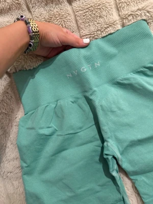 Turkosa träningstights-shorts från NVGTN - Säljer ett par turkosa seamless träningstights-shorts från NVGTN med hög midja och ribbad linning. De är stretchiga och sitter tight, perfekta för gymmet eller löpning. Snygg och sportig look med diskret logga framtill.