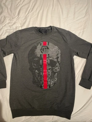 Philipp Plein grå tröja med döskalle - Snygg grå tröja från Philipp Plein med ett stort döskallemotiv i silver och svart på framsidan, samt en röd och svart rand i mitten. Axelpartiet har coolt texturerat mönster och på ryggen står det 'PLEIN' i rött. Helt ny och bra passform🌟