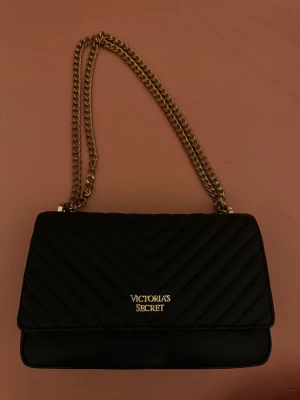 Svart quiltad axelväska Victoria's Secret - Snygg svart axelväska från Victoria's Secret med quiltad design och guldfärgad kedja. Väskan är i skinnimitation och har ett randigt foder på insidan. Perfekt storlek för det viktigaste och ger en lyxig känsla till din outfit. Den är köpt för från london för 90£ alltså ungefär 1100kr. En heldel färg på kjedan har försvunnit. !!PRIS KAN DISKUTERAS!!