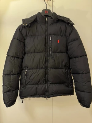 Svart pufferjacka Polo Ralph Lauren - Svart pufferjacka från Polo Ralph Lauren med röd logga på bröstet. Jackan har huva, flera fickor med dragkedja och quiltad design. Perfekt för kalla dagar och har en clean, sportig look. Materialet är syntet och dun, med långärmade ärmar och justerbara detaljer vid huvan.