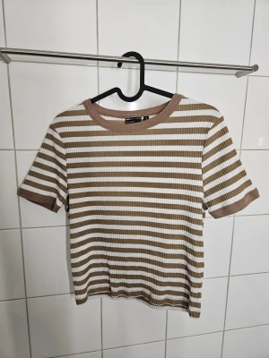 Randig ribbad t-shirt - Säljer en ribbad t-shirt från ASOS DESIGN med beige och vita horisontella ränder. T-shirten har korta ärmar, rund halsringning och är lite croppad. Den har en liten fläck på baksidan (se sista bilden).