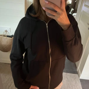 Svart hoodie med dragkedja från Lager 157 - Svart hoodie från Lager 157. Tröjan har dragkedja framtill, huva med snörning och två stora fickor. Tillverkad i mjuk bomullsmix, perfekt för chill dagar eller när du vill ha en enkel och stilren look. Storlek xs men lite längre och lösare passform. 