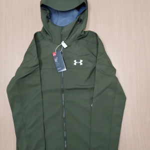 Under armour jacka  - Under Armour Softshell-jacka – Grön Snygg och funktionell softshell-jacka från Under Armour i mörkgrön färg. Jackan är mycket bekväm och har en normal passform, perfekt både för vardag och träning utomhus. Den ser ut som storlek L men motsvarar snarare en större M – alltså lite rymligare än M men inte riktigt L.  Skick: Mycket bra, nästan nyskick. Endast sparsamt använd, inga synliga defekter, hål eller fläckar.  Material & funktion: Tillverkad i vindtätt och vattenavvisande softshell-material. 