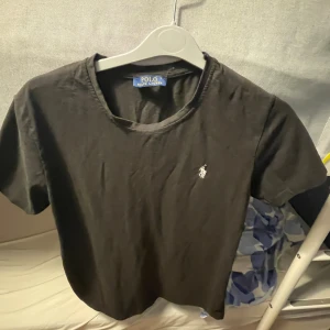 Svart t-shirt från Polo Ralph Lauren - Svart t-shirt från Polo Ralph Lauren med klassisk rund hals och broderad vit logga på bröstet. Enkel och stilren design i mjuk bomull, perfekt till jeans eller shorts. Skön passform och tidlös look.