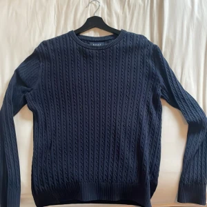Mörkblå kabelstickad tröja från Riley - Kabelstickad mörkblå tröja från Riley i klassisk modell med rund halsringning och ribbade muddar. Tröjan har lång ärm och ett snyggt strukturmönster som ger en stilren och tidlös look. Nyskick och sitter väldigt bra!🌟🌟