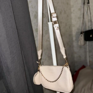 Axelväska med kedjedetalj - Snygg axelväska i croco-mönstrat syntetmaterial med bred justerbar axelrem och guldfärgad kedja framtill. Väskan har dragkedja upptill och en liten rund miniväska på sidan, perfekt för småprylar. Stilren och trendig accessoar till din outfit.