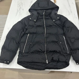 Svart pufferjacka från BOSS med huva - Svart pufferjacka från BOSS med diskret mönster, avtagbar huva och silvriga dragkedjor. Jackan har långa ärmar, två stora fickor med dragkedja och tryckknappar vid ärmsluten. Perfekt för kalla dagar och riktigt snygg streetstil.