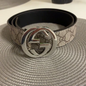 GG Supreme bälte med silverdetalj - Snyggt bälte med klassiskt GG Supreme-mönster i beige och brunt, samt en stor silverfärgad dubbel-G-spänne. Baksidan är svart skinn. Perfekt accessoar för att lyfta din outfit med en lyxig touch.