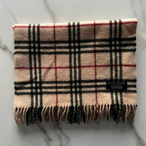 Burberry halsduk beige - Klassisk halsduk från Burberry i beige med rutigt mönster i svart, vitt och rött. Gjord i 100% lammull och har snygga fransar i ändarna. Perfekt accessoar för att lyfta din outfit med en tidlös brittisk touch. En av Burberrys nyare modeller, har en liten annan färg en den vanliga beige halsduken.