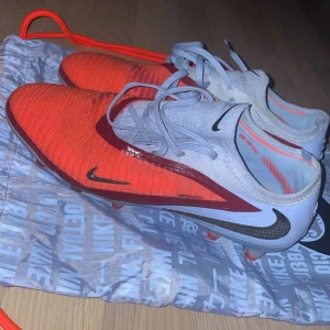 Nike phantom gx 6 elite/ Ag. Treadar mot nya f50s i storlek 42 2/3 Ele 43 - Säljer ett par Nike fotbollsskor med snygg design i orange och ljusgrått. Skorna har snörning, svarta Nike-loggor och röda detaljer längs sidan. Ovandelen är i syntetmaterial och sulan är anpassad för spel på gräs. Perfekta för dig som vill sticka ut på planen!
