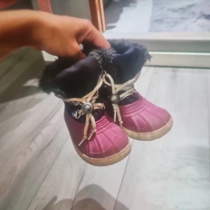 Vinröda vinterboots med fuskpäls - Säljer ett par vinröda boots med svart fuskpäls runt skaftet och beige snörning med coola detaljer. Skorna har en platt sula och är fodrade för att hålla värmen under kalla dagar. Perfekta för vintern och riktigt snygga till jeans eller leggings. Skor för barn!!