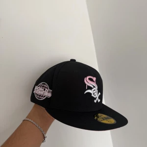 New Era keps - Säljer en svart 59FIFTY keps från New Era med White Sox-logga i vitt och rosa framtill. Kepsen har broderad World Series-patch på sidan och en platt skärm med rosa undersida. Tillverkad i bomull och polyester, perfekt för dig som gillar sportig streetstyle.