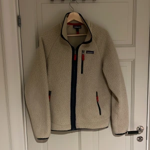 Beige teddyjacka från Patagonia M - Beige teddyjacka från Patagonia i mysig fleece med marinblå detaljer och röda dragkedjor. Jackan har hög krage, tre fickor med dragkedja och klassisk Patagonia-logga på bröstet. Perfekt för kalla dagar och chill stil.