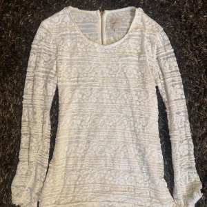 Vit spetsblus med dragkedja  - Säljer en vit långärmad blus från Carela F med vackert blommigt spetsmönster och rund halsringning. Blusen har dragkedja i ryggen och är perfekt för dig som gillar romantisk och feminin stil.