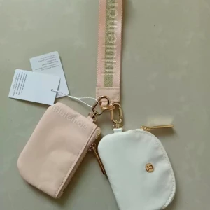 Lululemon plånbok duo i rosa och vitt - Säljer ett set med två små plånböcker från Lululemon, en i ljusrosa och en i vit, båda med silverfärgade dragkedjor och logga. De sitter ihop med en bred, rosa handledsrem och metallkedjor. Perfekt för kort, mynt eller småsaker.
