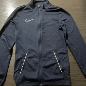 Marinblå Nike träningsjacka XS och marinblå Nike träningsbyxor XS - Nike Dri-Fit träningsjacka i marinblått med hel dragkedja framtill och vita detaljer vid fickorna. Jackan har lång ärm, hög krage och klassisk Nike-logga på bröstet. Perfekt för sport eller chill dagar. Precis samma ingår för byxorna samma storlek och allting det är ett matchande sett som köptes tillsammans. Mycket fint skick i båda plaggen