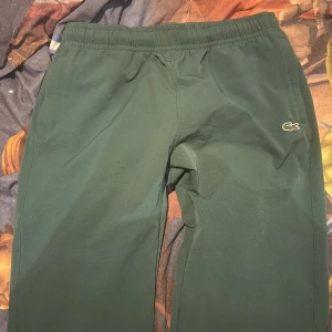 Gröna mjukisbyxor från Lacoste Sport - Mörkgröna mjukisbyxor från Lacoste Sport med elastisk midja och vit snörning. Byxorna har dragkedjeförsedda fickor och klassisk Lacoste-logga broderad på benet. Perfekta för chill eller träning, riktigt sköna och stilrena.