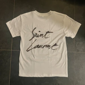 Vit t-shirt med tryck - Vit t-shirt från Saint Laurent med stort svart handskrivet logotyptryck på ryggen. Klassisk passform och rund hals. Perfekt för dig som vill ha en clean look med exklusiv känsla.