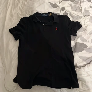Svart pikétröja Polo Ralph Lauren - Klassisk svart pikétröja från Polo Ralph Lauren i slim fit-modell. Tröjan har korta ärmar, krage och knäppning framtill samt den ikoniska röda broderade loggan på bröstet. Perfekt för en clean och stilren look.