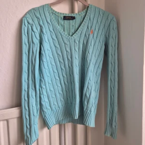 tröja Polo Ralph Lauren -  ljusblå kabelstickad tröja från Polo Ralph Lauren med v-ringning och broderad logga i orange på bröstet. 