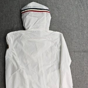 Vit Moncler-liknande dunjacka med huva och dragkedja (oanvänd och oöppnad helt ny) - Helt Ny, Stilren oanvänd, och oöppnad i en Moncler-liknande förpackning, är en vit Moncler-liknande dunjacka med huva och dragsko, hel dragkedja framtill och två fickor med knappstängning. Jackan har en enkel design med diskreta detaljer och en liten randig detalj på huvan baktill. Perfekt för lager-på-lager och snygg till streetwear.