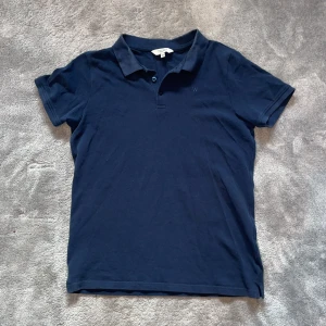 Marinblå pikétröja från Wayfarer Young - Snygg marinblå pikétröja från Wayfarer Young med klassisk krage och knäppning framtill. Tröjan är i bomull och har korta ärmar, perfekt för en stilren och avslappnad look. Passar dig som gillar enkel och tidlös stil.