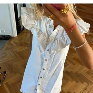Vit volangklänning från Zara XS - Supersöt vit kortklänning från Zara med volangdetaljer vid axlarna och knappar hela vägen framtill. Klänningen har en rak passform och är ärmlös med markerad krage. Perfekt för dig som gillar en clean och trendig look.