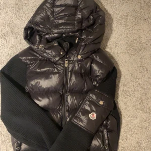 Svart Moncler cardiganpufferjacka med huva - Säljer en svart pufferjacka från Moncler med glansig finish, stickade ärmar och avtagbar huva. Jackan har dragkedja framtill, ribbade muddar och Moncler-logga på ärmen. Perfekt för kalla dagar och riktigt snygg till streetwear.
