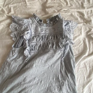 Ljusblå volangklänning Holly & Whyte - Supersöt ljusblå klänning från Holly & Whyte med volangdetaljer och broderad spets upptill. Klänningen har ärmlös design med romantiska volanger på axlarna och rynkade partier framtill. Perfekt för sommarens alla tillfällen!