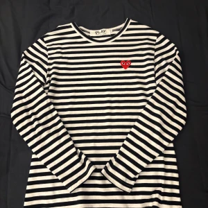 Randig långärmad tröja Comme des Garçons - Säljer en svartvit randig långärmad tröja från Comme des Garçons Play. Tröjan har ett ikoniskt rött hjärta med ögon broderat på bröstet och är tillverkad i mjuk bomull. Perfekt för dig som gillar stilrena och trendiga plagg. Storlek L