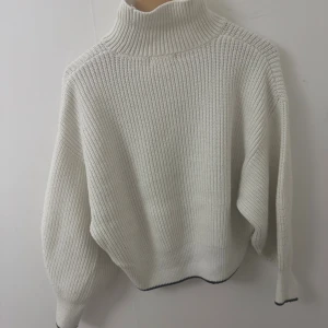 Vit stickad polotröja med ribbad struktur - Vit stickad polotröja med hög krage och ribbad struktur. Tröjan har en loose passform och långa ärmar, perfekt för kyliga dagar. Diskret mörk kant längst ner på ärmar och nederkant ger en snygg detalj.