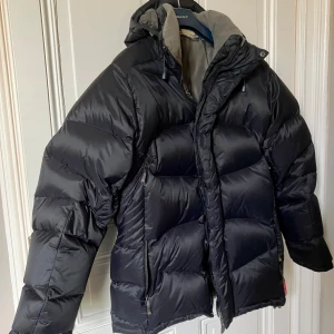 Svart dunjacka från Salomon L - Säljer en svart dunjacka från Salomon i storlek L. Jackan är rejält vadderad, har huva och dragkedja framtill samt två sidofickor med dragkedja. Perfekt för kalla dagar och har en clean, sportig look.