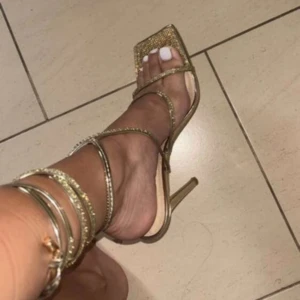 Glittriga guldiga  klackskor med snörning - Säljer ett par glittriga klackskor i guld med tunna remmar och snörning runt vristen. Skorna har fyrkantig tå och hög klack, perfekt för dig som vill sticka ut. Materialet är syntet med glittriga detaljer som fångar ljuset. Riktigt snygga till en partylook!