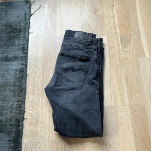 Svarta jeans från Nudie, 30/30 - Säljer ett par svarta jeans från Nudie Jeans i storlek 30/30. Jeansen har en snygg tvättad look med slitningar. De är raka i modellen. Perfekta för dig som gillar en avslappnad stil. Det är bara att höra av dig om du är intresserad!